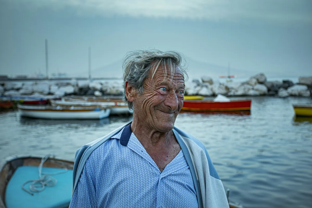A weathered older man smiling by a small harbor